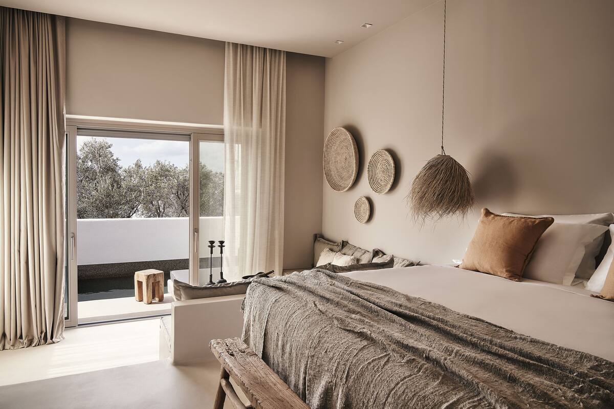 A bedroom with a large bed, a window with curtains, a view of trees, and a small wooden stool.