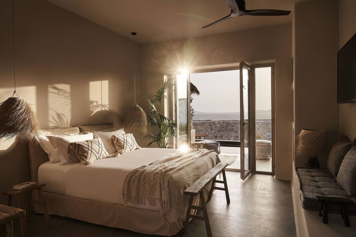 A bedroom with a large bed, a bench, a sofa, and a view of the outdoors.