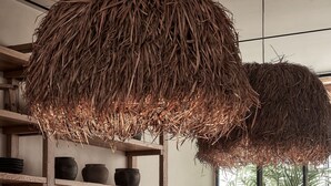 Ein Holztisch mit einer gewebten Korb-Mitteldeko, rustikalen Kerzenhaltern und Blick auf Palmen durch ein großes Fenster.