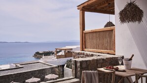 Eine Terrasse mit Steinmauer, Holzmöbeln, einem Tisch mit Flasche und Gläsern sowie Blick auf das Meer und das entfernte Land.