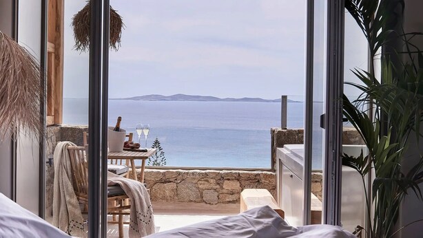 Ein Schlafzimmer mit großem Fenster und Blick auf die Küste, einem Holztisch mit Flasche und Gläsern sowie einer hängenden Korbin.