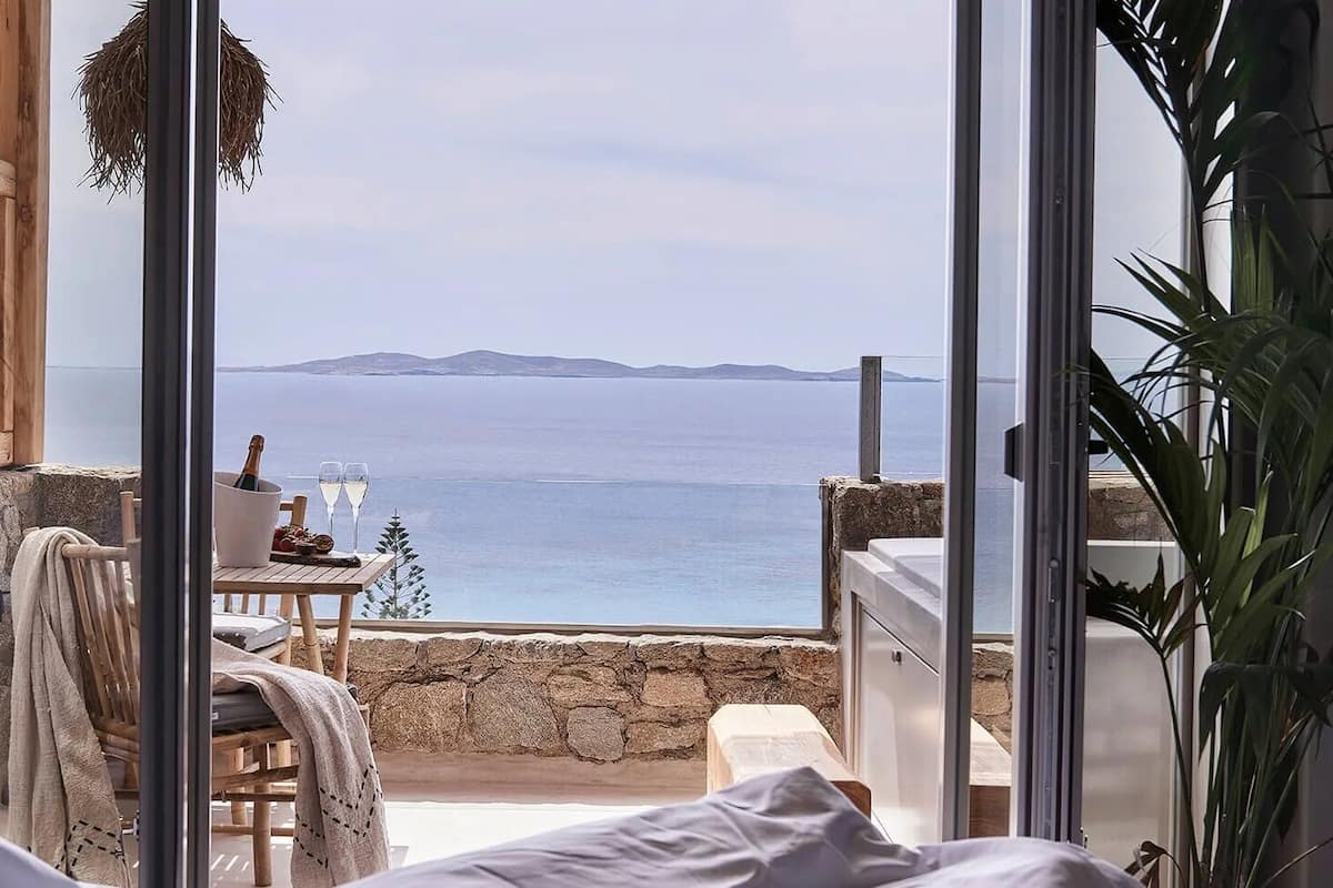 A bedroom with a large window overlooking a coastal view, a wooden table with a bottle and glasses, and a hanging basket.