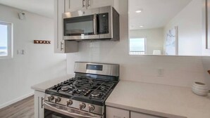 A modern kitchen with stainless steel appliances, white cabinetry, and a large window providing natural light. - The Buccaneer House - Oceanfront (Oceanside)
