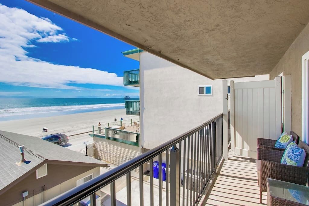 A balcony with a view of the beach, a railing, and a wicker sofa with cushions.