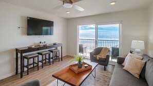 A modern living room with a wooden coffee table, a sofa, a TV mounted on the wall, and a balcony with a view. - The Buccaneer House - Oceanfront (Oceanside)