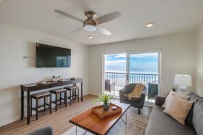 A modern living room with a wooden coffee table, a sofa, a TV mounted on the wall, and a balcony with a view. - The Buccaneer House - Oceanfront (Oceanside)