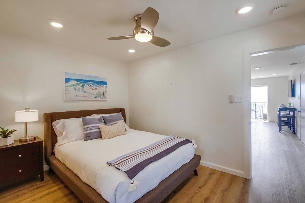 A bedroom with a bed, a ceiling fan, a nightstand with a lamp, and a painting on the wall.