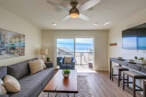 Un salon moderne avec un canapé, une table basse et un téléviseur. Il y a un balcon avec vue sur l’océan.