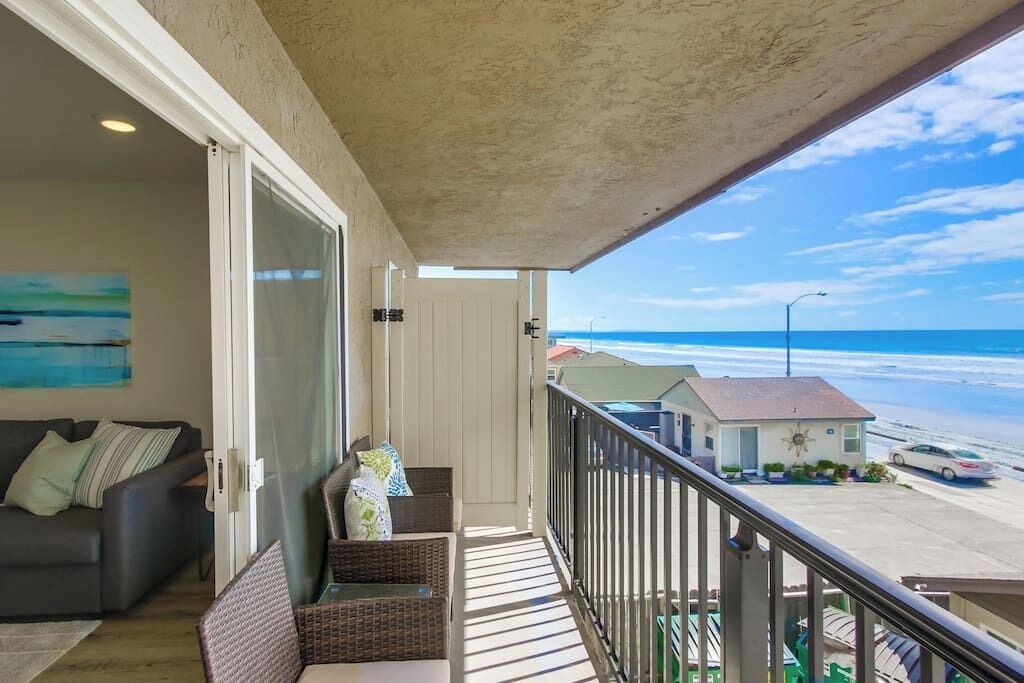 A balcony with a view of the beach, a sofa, wicker chairs, and a small house in the background.