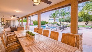 A covered patio with a wooden table and chairs, a ceiling fan, and a view of a swimming pool and greenery. - Centrally located estate in Scottsdale with a heated pool, game room, and more! (Scottsdale)