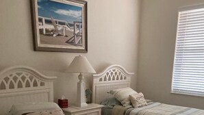 Un dormitorio con dos camas, una mesita de noche con lámpara, un cuadro enmarcado en la pared y una ventana con persianas.