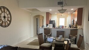 Una sala de estar con un sofá marrón, una mesa de comedor con sillas, y una cocina con refrigerador, microondas y armarios.