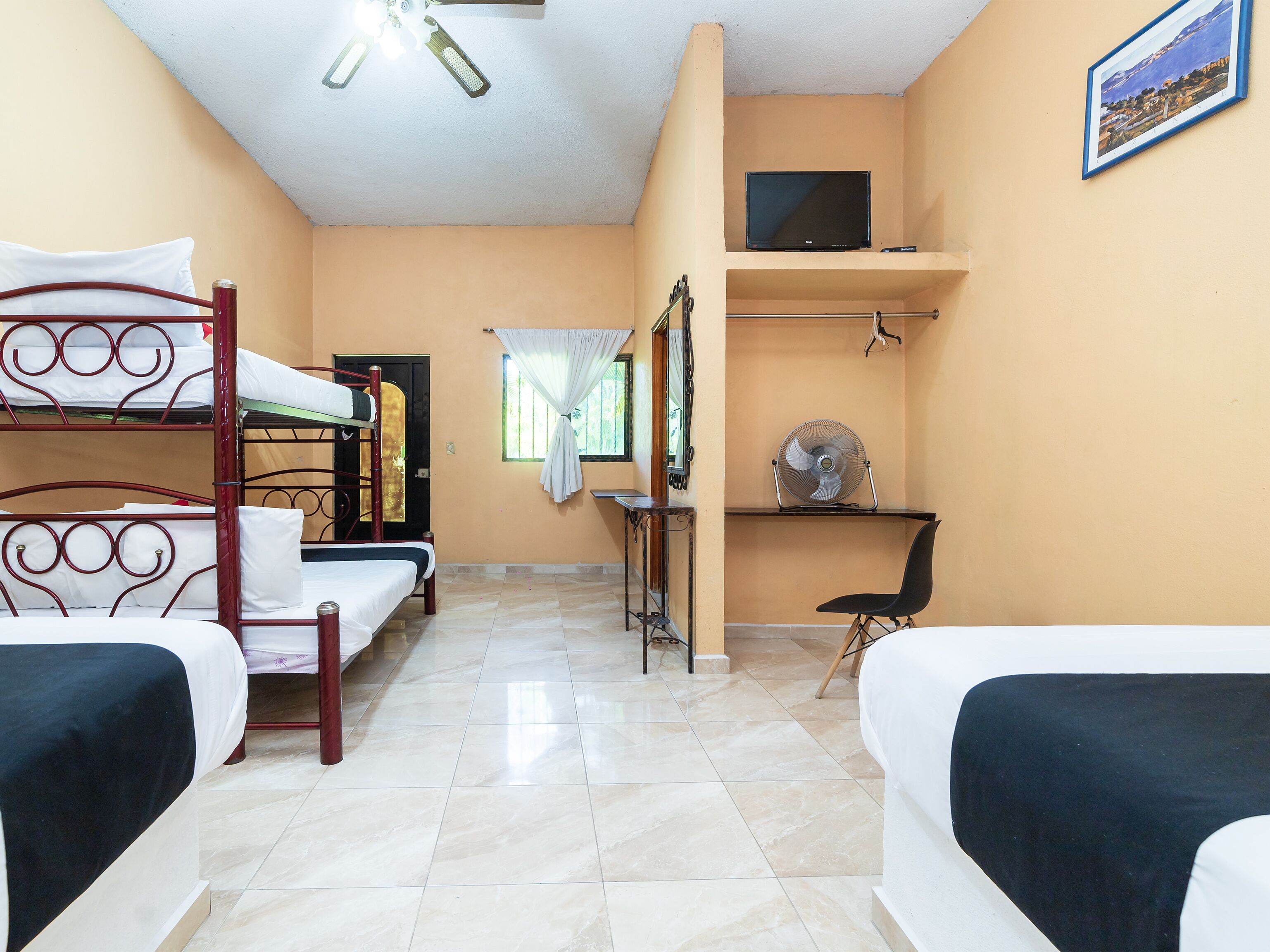 A dormitory room with bunk beds, a desk, a chair, a fan, a television, and a framed picture on the wall.