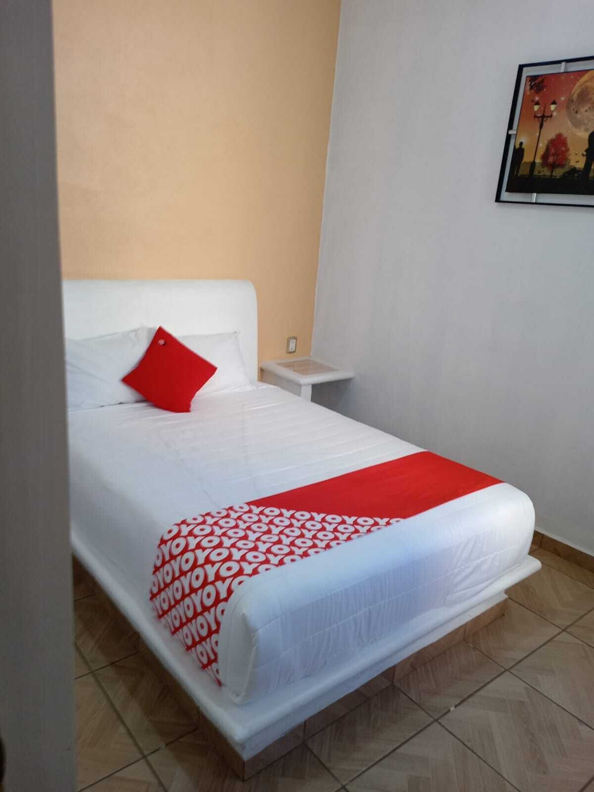 Una cama bien hecha con una colcha blanca y roja, un cojín rojo y una mesita de noche con una lámpara.