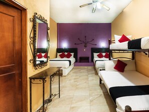 Habitación de hotel con dos camas individuales, una cama doble, un escritorio de madera, un espejo y un ventilador de techo.