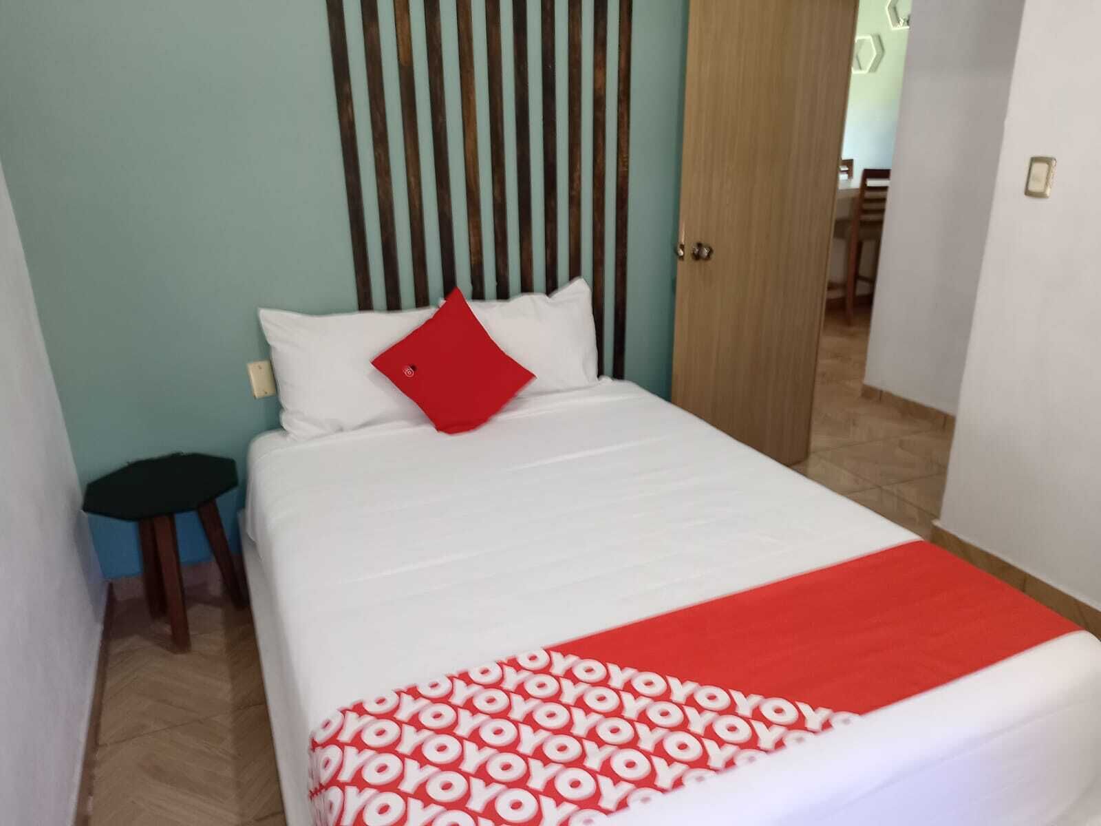 Habitación de hotel con una cama, una almohada roja, una colcha blanca y una manta estampada en rojo y blanco.