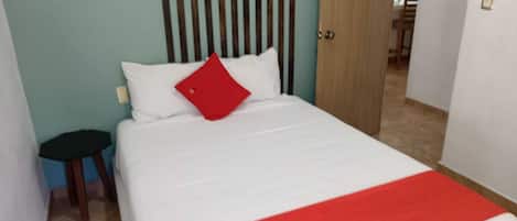 A hotel room with a bed, a red pillow, a white bedspread, and a red and white patterned throw.