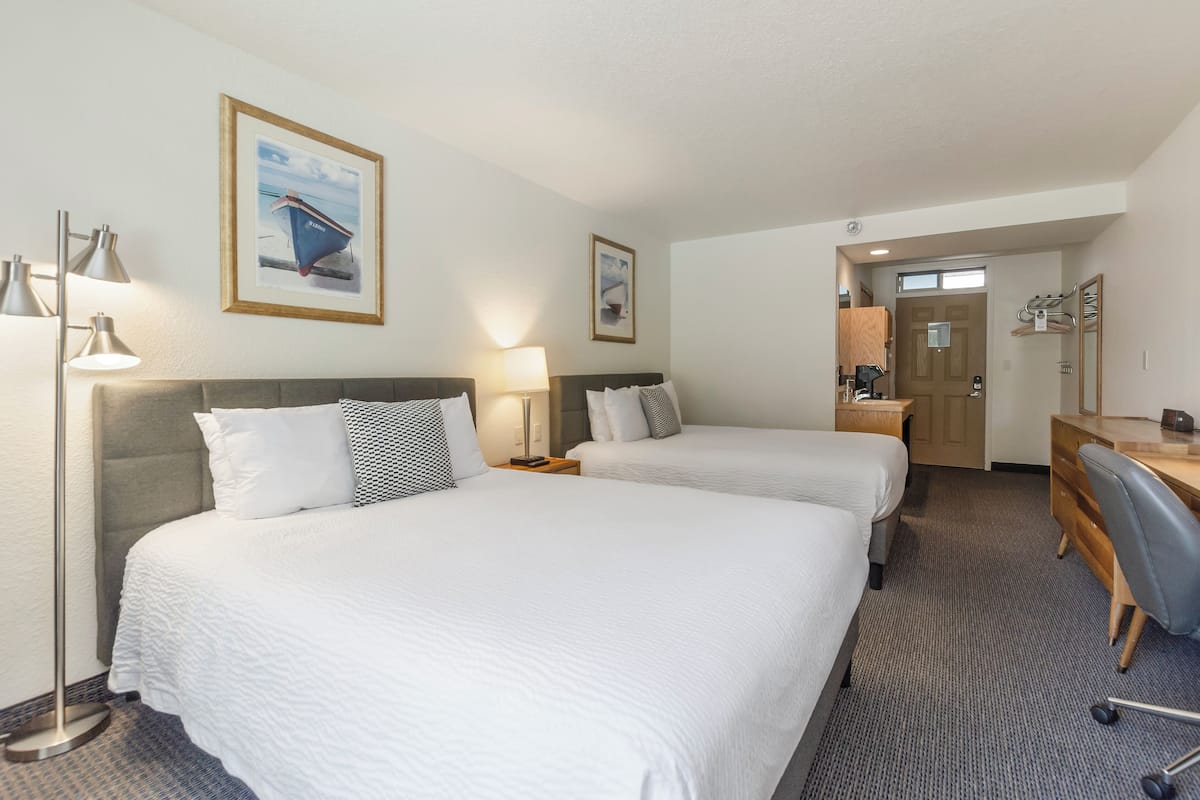 Une chambre d’hôtel avec deux lits, un bureau, une chaise et une salle de bain visible à travers une porte ouverte.