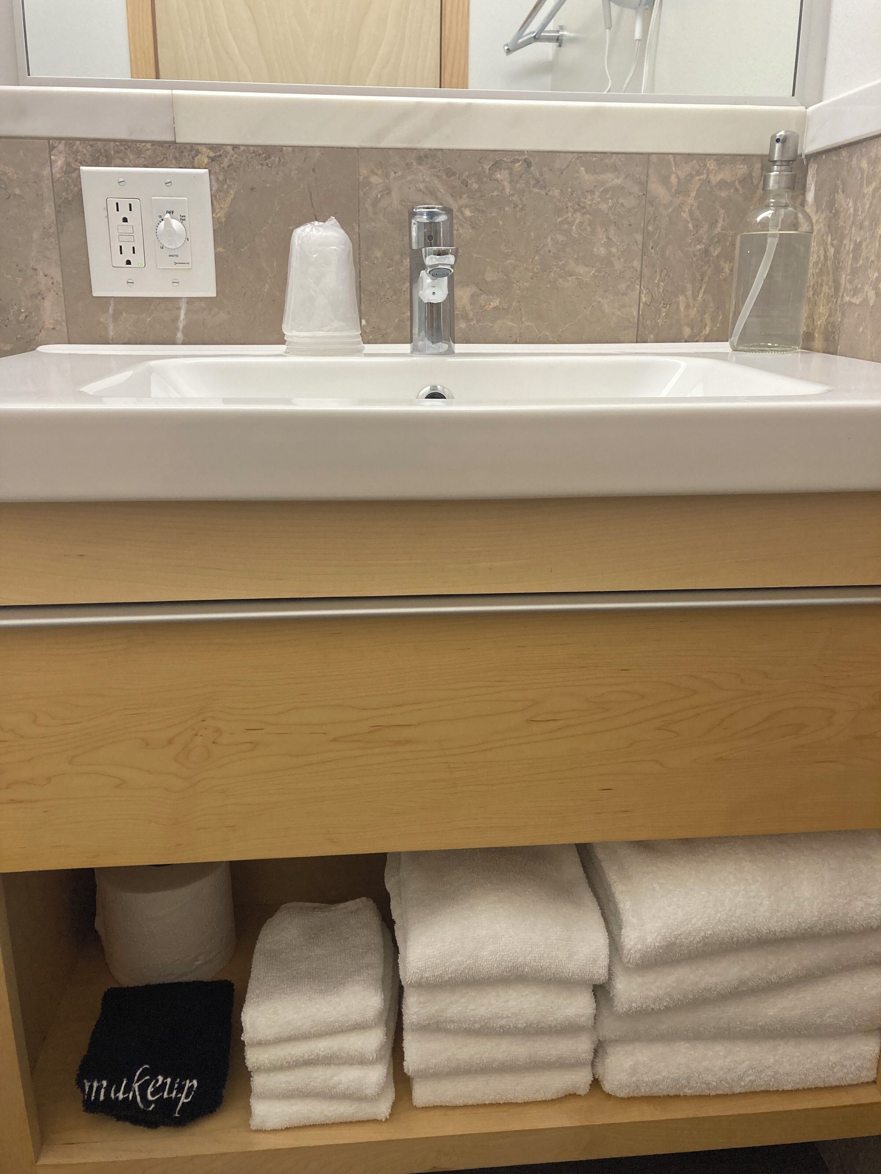 Une salle de bain avec un lavabo blanc, un miroir, un porte-serviettes avec des serviettes blanches et une étagère sur laquelle se trouve une serviette noire étiquetée « maquillage ».