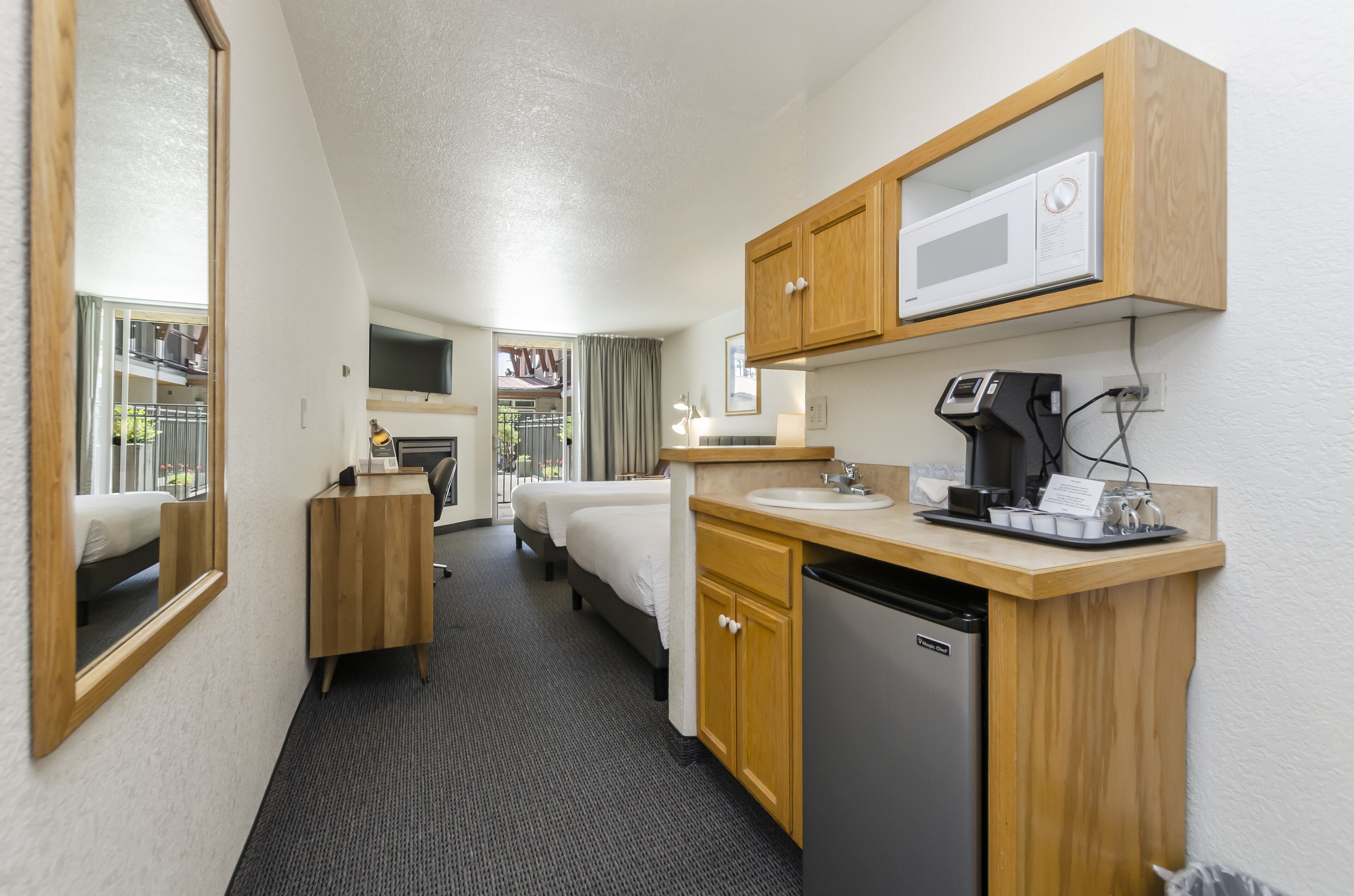 A compact hotel room with a kitchenette, a bed, a desk, and a mirror.