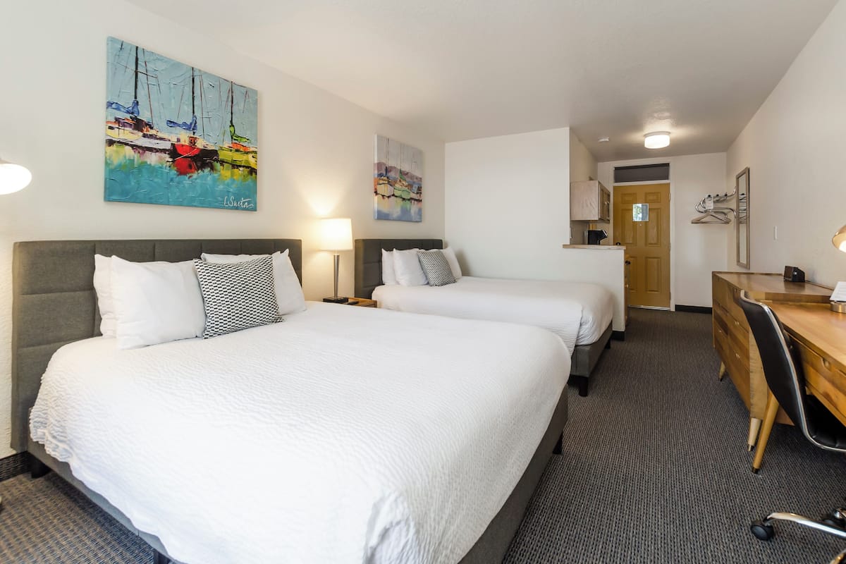 Une chambre d’hôtel avec deux lits, un bureau et une chaise. Les murs sont ornés de tableaux et une porte donne accès à la salle de bain.