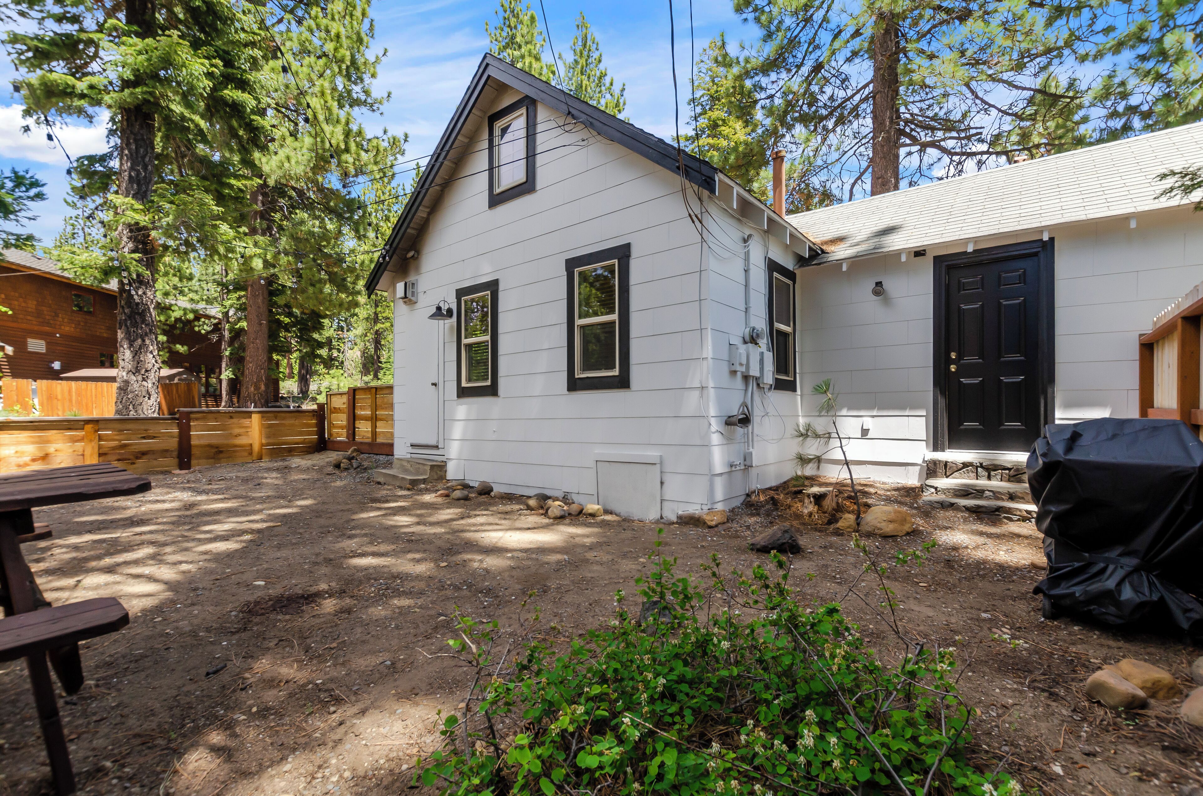A single-story house with a white exterior and black trim, surrounded by a wooden deck and a dirt yard with rocks and greenery.