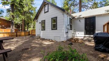 A single-story house with a white exterior and black trim, surrounded by a wooden deck and a dirt yard with rocks and greenery.