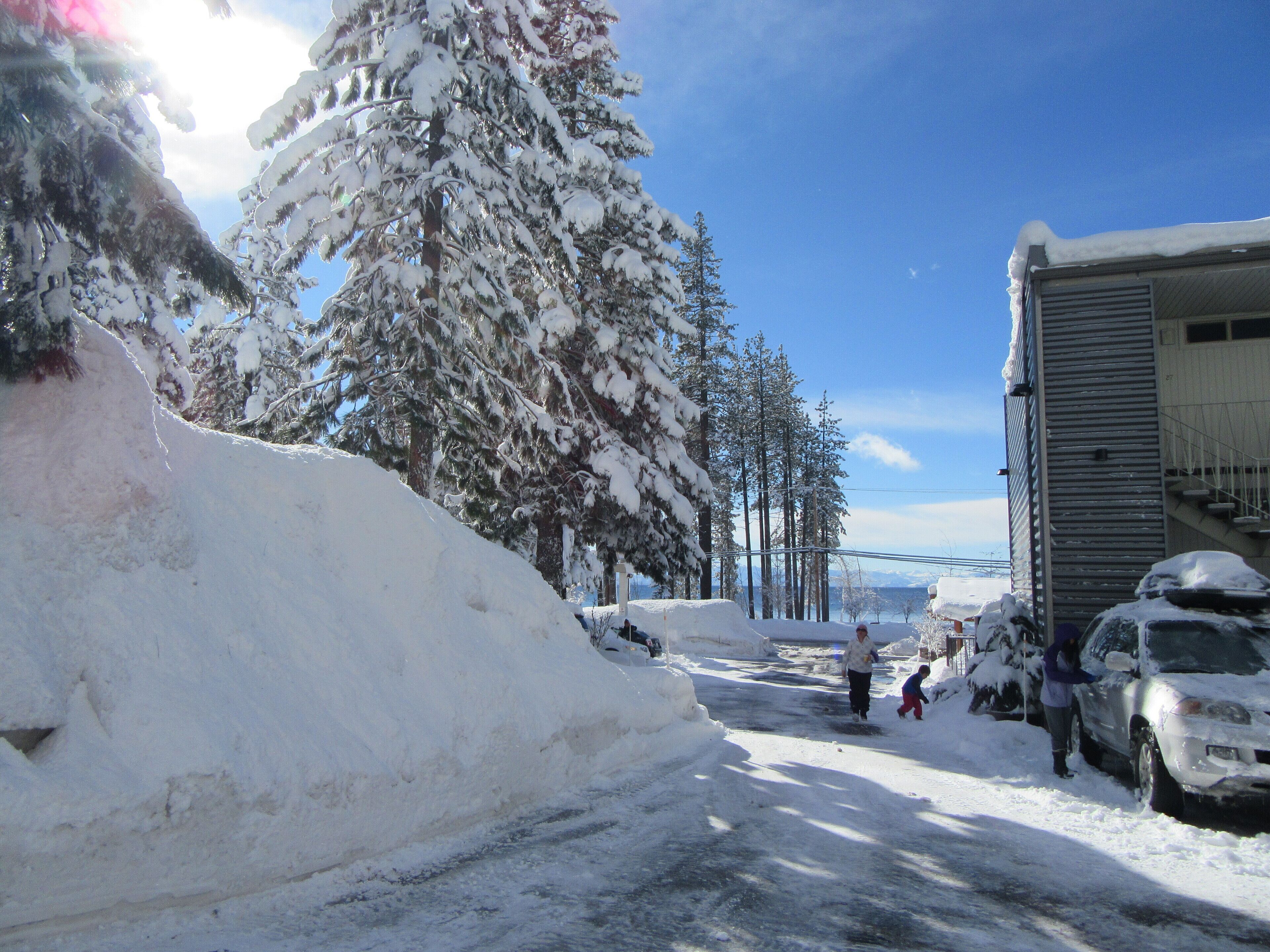 A snowy street with a large snowbank on the left, a building on the right, and a clear blue sky.