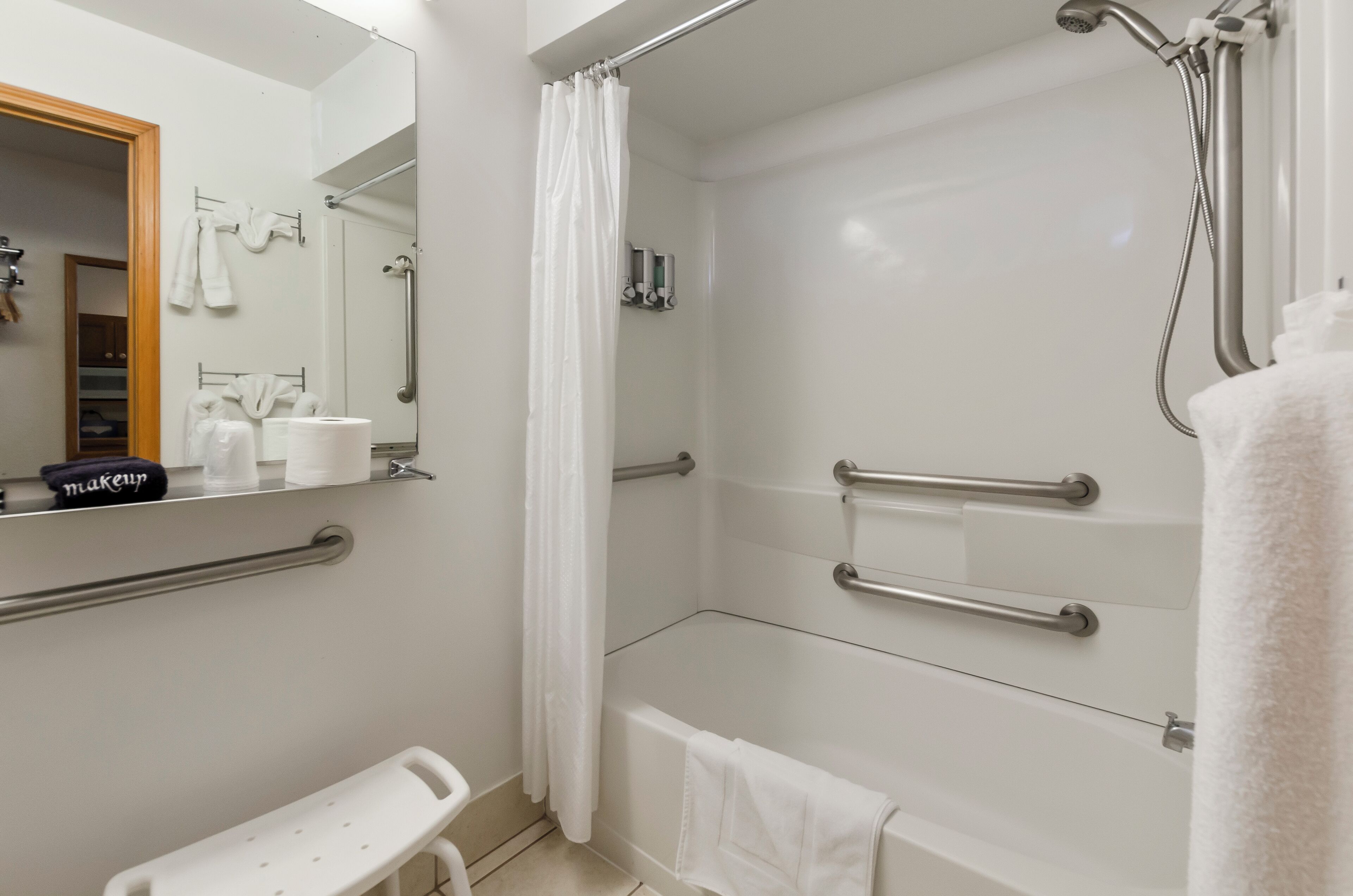 Un baño moderno con cabina de ducha, barras de apoyo, un banco y un espejo con toallero.