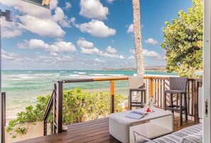 Une terrasse en bois avec un canapé blanc et deux fauteuils en osier, donnant sur une plage et l’océan.