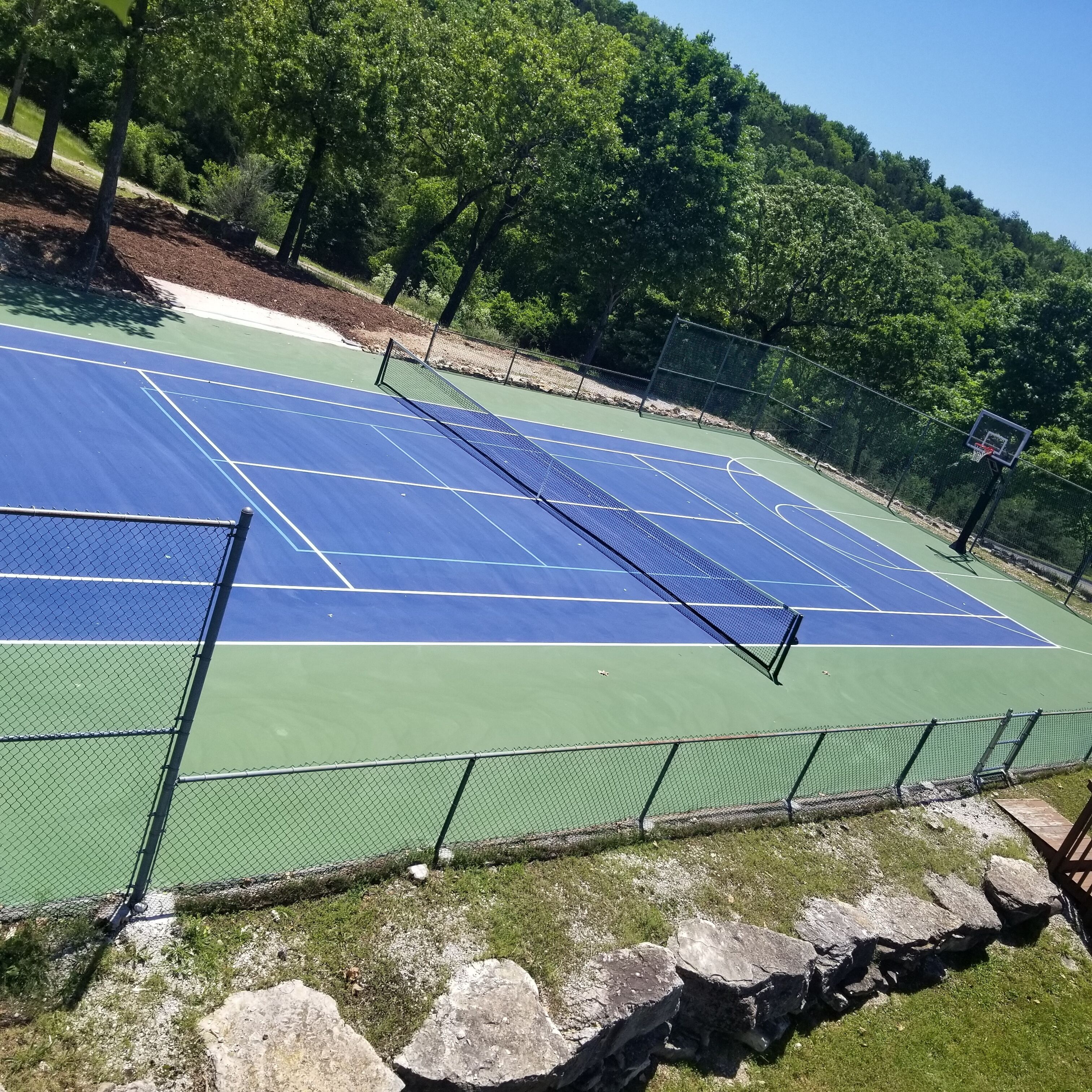 Een tennisbaan met een net, omringd door een hek en bomen.