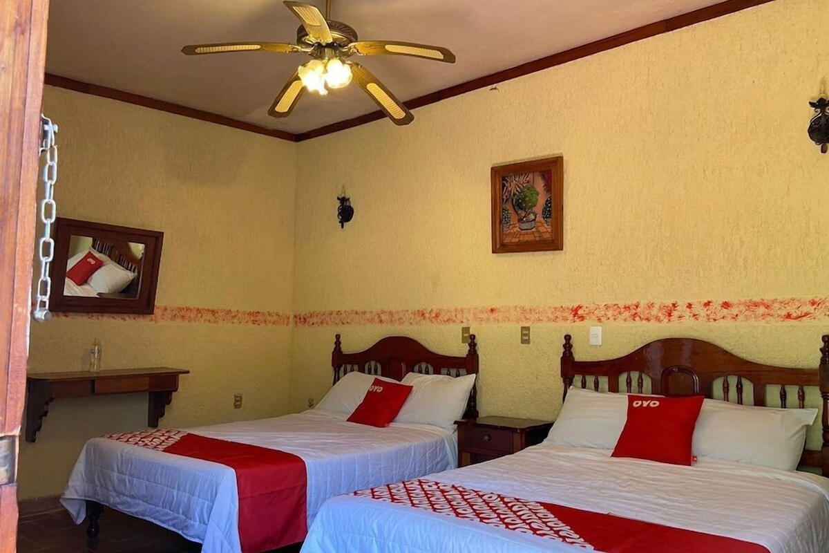 A hotel room with two beds, a ceiling fan, a small table, and a wall-mounted light fixture.