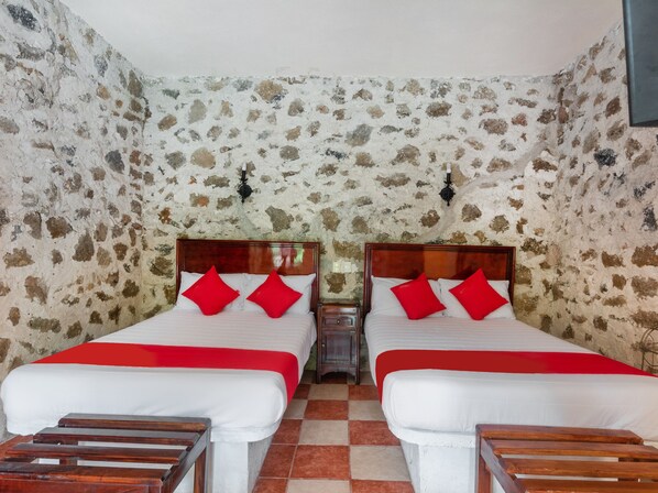 Una habitación con dos camas, cada una con almohadas rojas y una colcha blanca. Las paredes tienen un patrón con textura de piedra.