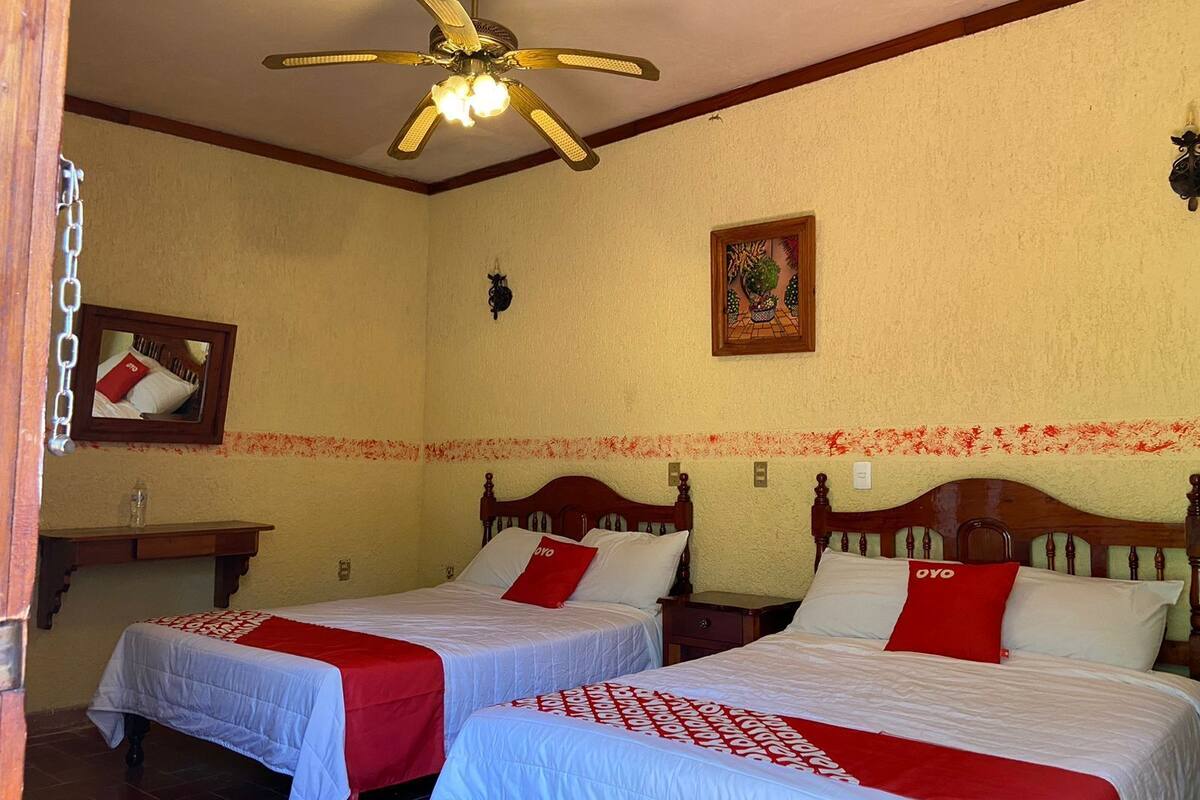 A hotel room with two beds, a ceiling fan, a mirror, a small table, and a decorative wall piece.