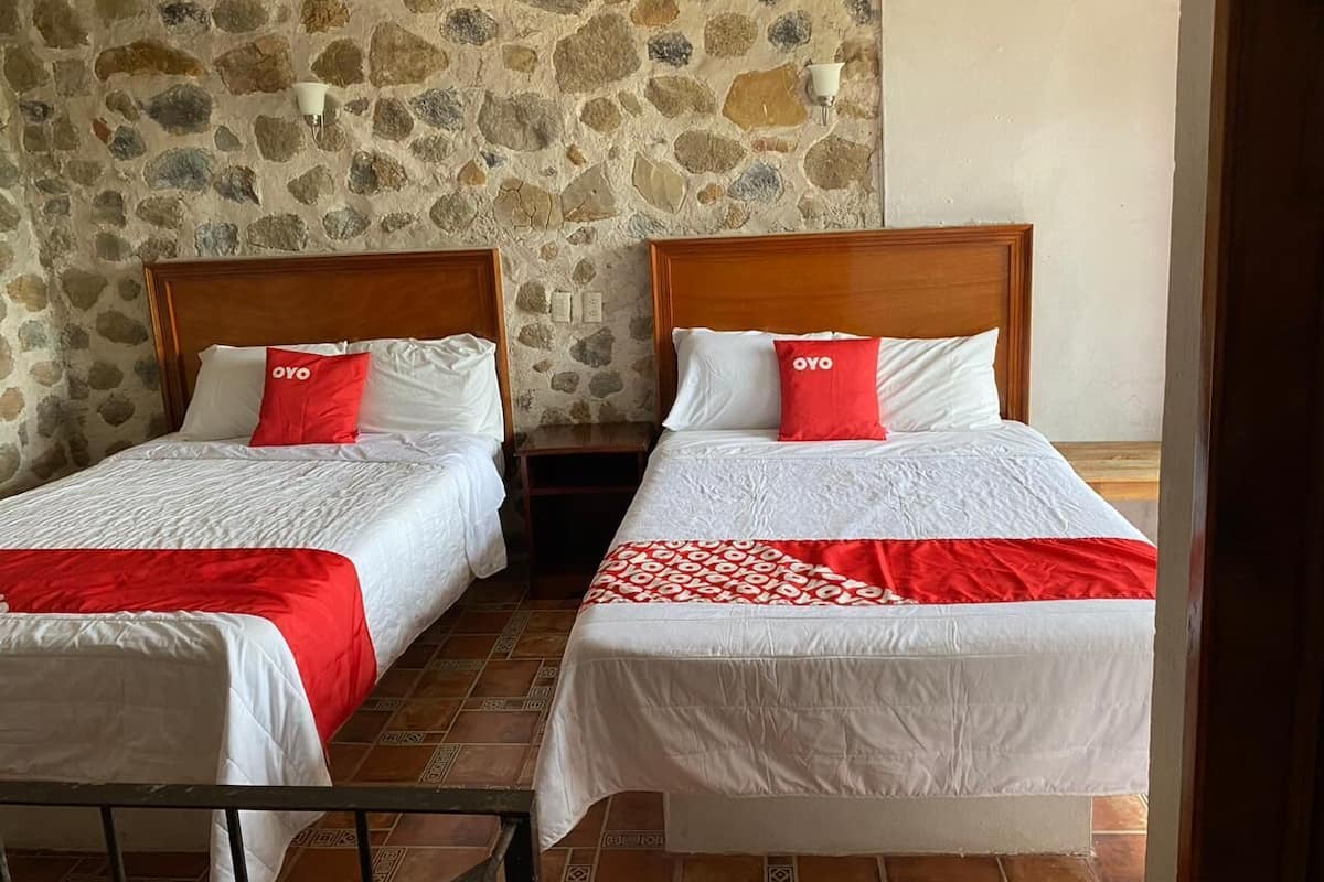 Two single beds with wooden headboards, white bedding, and red pillows with 'OYO' branding. The room has a stone wall and tiled floor.