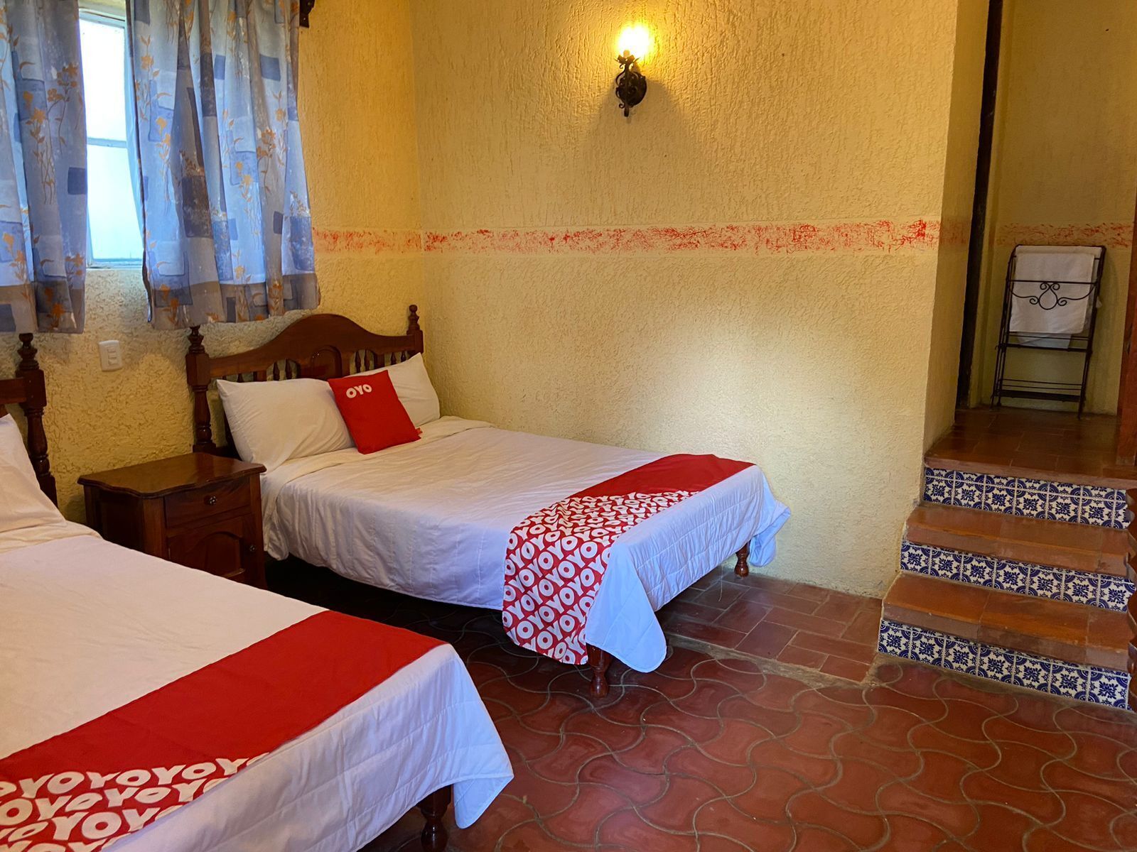 Habitación de hotel con dos camas, una mesita de noche, una lámpara fijada a la pared y una escalera con azulejos estampados.