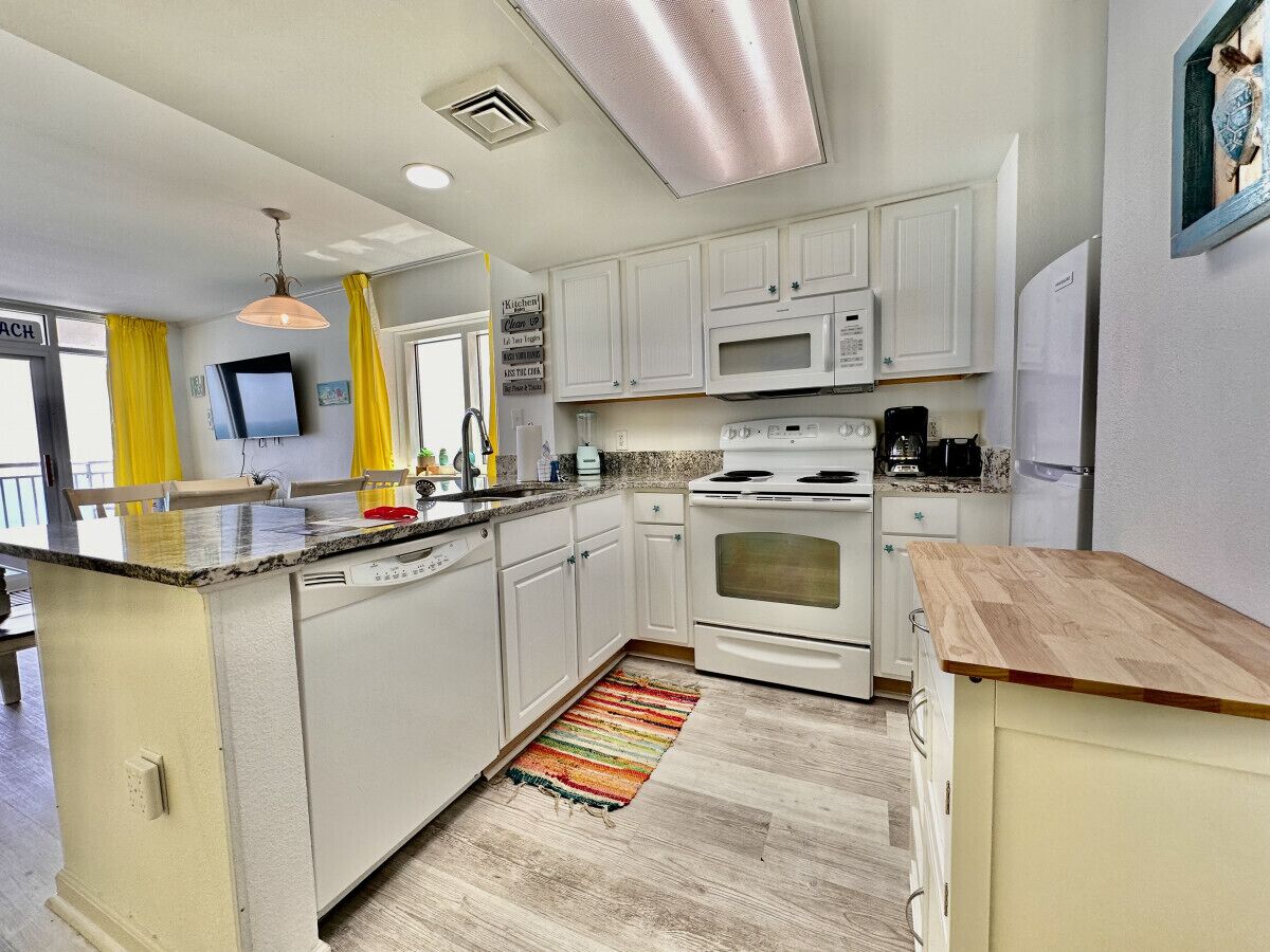 A compact kitchen with white cabinets, a microwave, an oven, and a dishwasher. A colorful rug is on the floor, and a wooden countertop is visible.