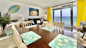 A spacious living room with a large wooden table, comfortable seating, a sofa, and a view of the ocean through large windows.