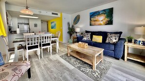 A modern living room with a blue sofa, wooden coffee table, and a dining area with white chairs.