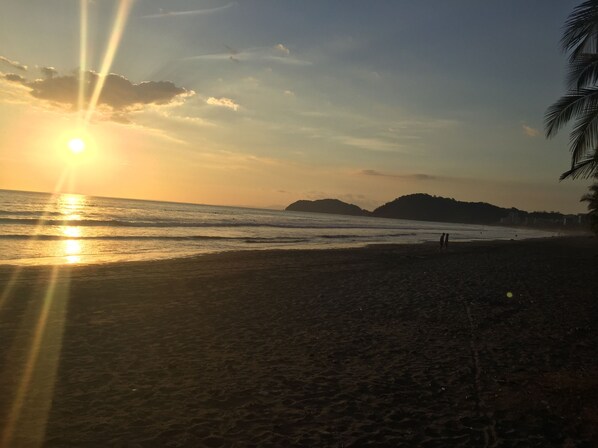 Uma praia ao pôr do sol, com o sol baixo no horizonte, mar calmo e colinas ao longe.
