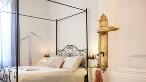 A four-poster bed with a white bedspread and two pillows, a brass door handle, and a small table with a lamp beside the bed. - Luxury Designer Rome  — St. Peter Dome View (Roma)
