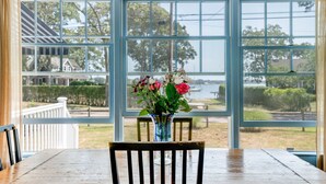 A dining area with a table and chairs, a vase with flowers, and a view of a lake and trees through large windows. - Lovely Shelter Island cottage with panoramic bay views, secluded, screened... (Shelter Island)