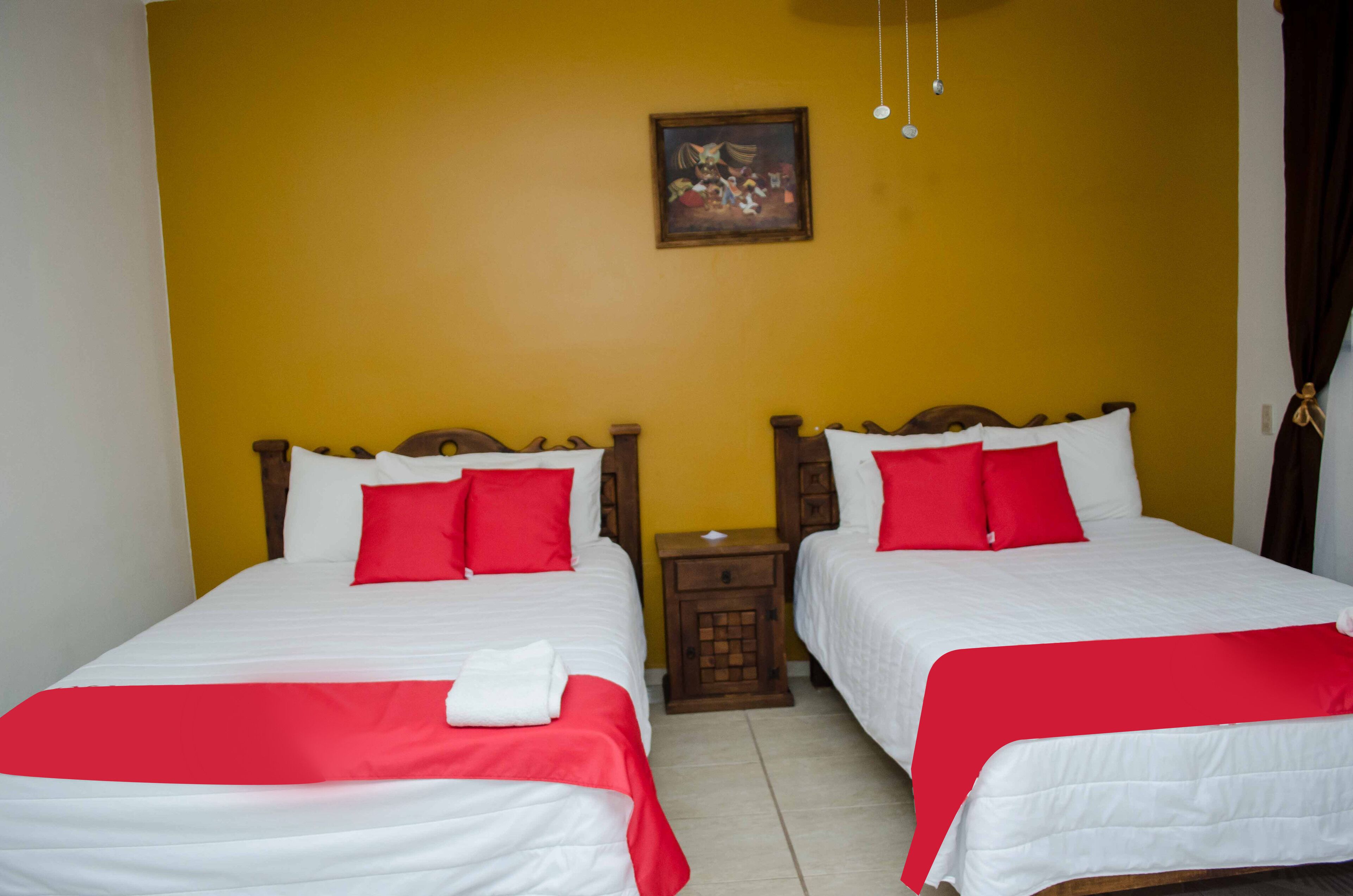 Une chambre d’hôtel avec deux lits, chacun agrémenté de draps blancs et de touches de couleur rouge. Une petite table de chevet en bois est placée entre les lits, et un tableau est accroché au mur jaune, juste au-dessus.