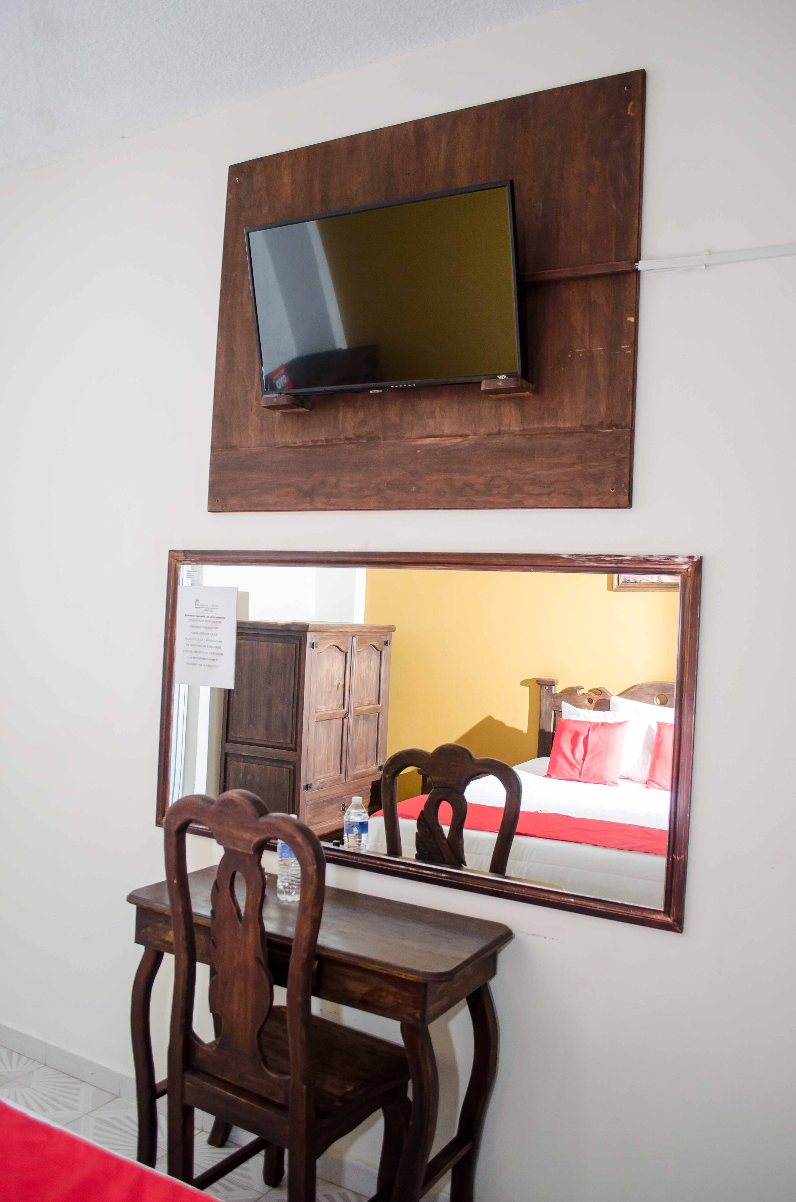 Une pièce dotée d’un téléviseur fixé au mur, d’une table à manger en bois avec des chaises, et d’un grand miroir qui reflète l’intérieur de la pièce.