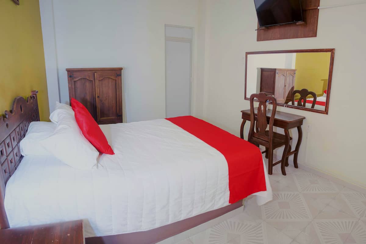 Una cama bien hecha con una almohada roja decorativa, un armario de madera, una mesita con una silla y un espejo en la pared.