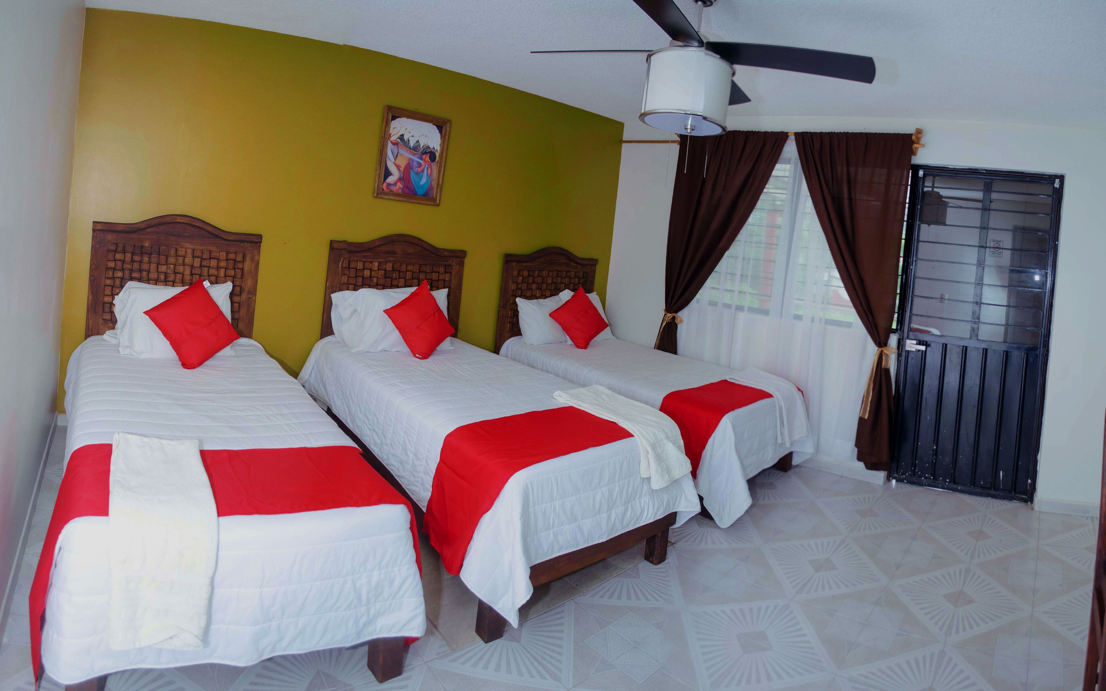 A hotel room with two single beds, a ceiling fan, a painting on the wall, and a window with curtains.