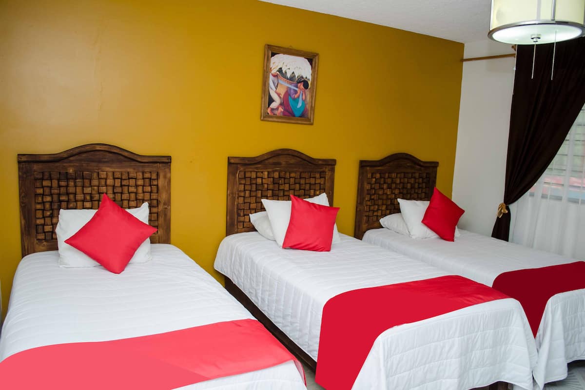 Habitación de hotel con tres camas individuales, cada una con cabecero de madera y ropa de cama blanca. En la pared amarilla, sobre las camas, hay un cuadro.