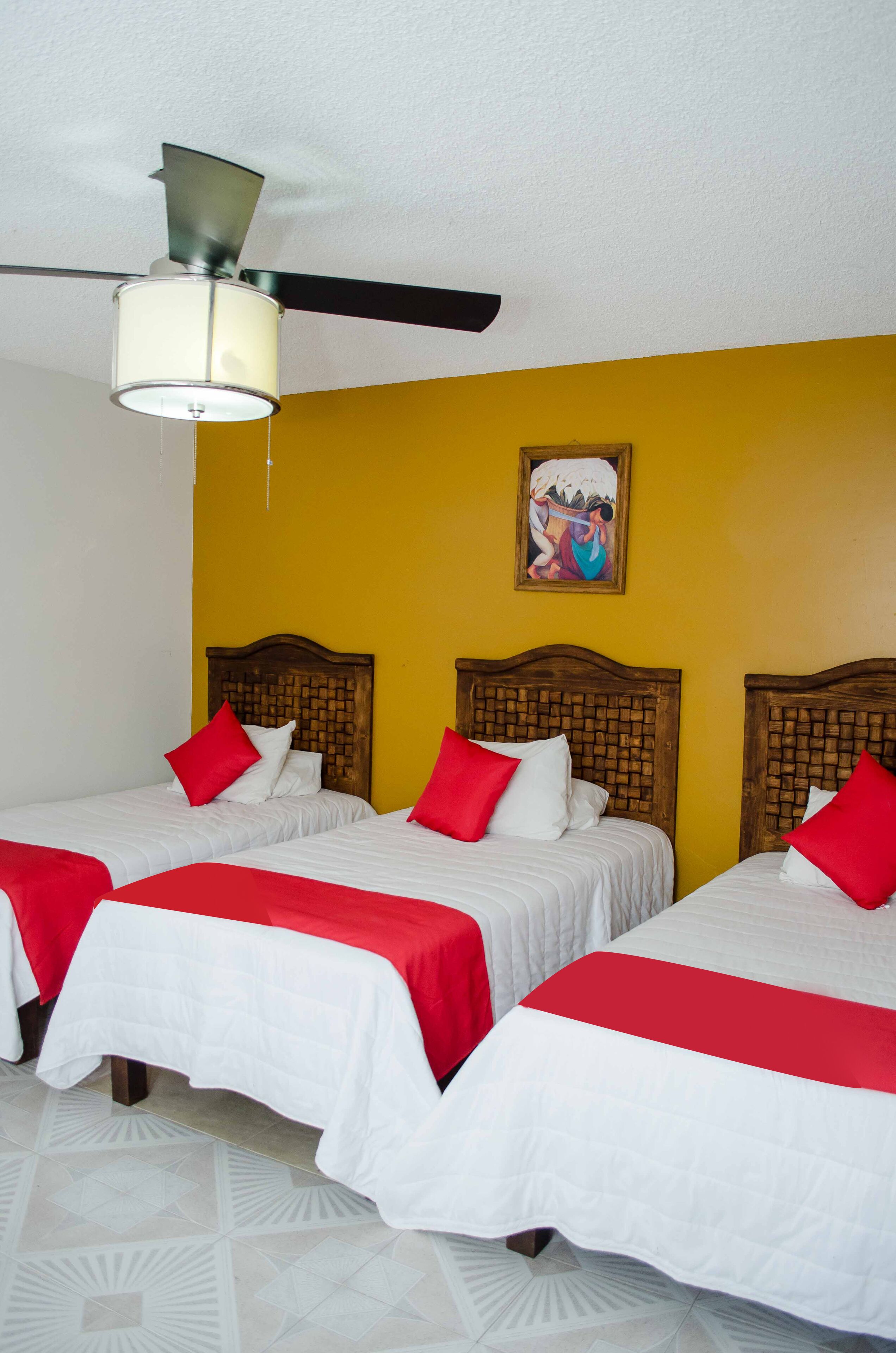 A hotel room with two beds, each with a wooden headboard, white bedding, and red accents. A ceiling fan is visible, and a painting hangs on the yellow wall.