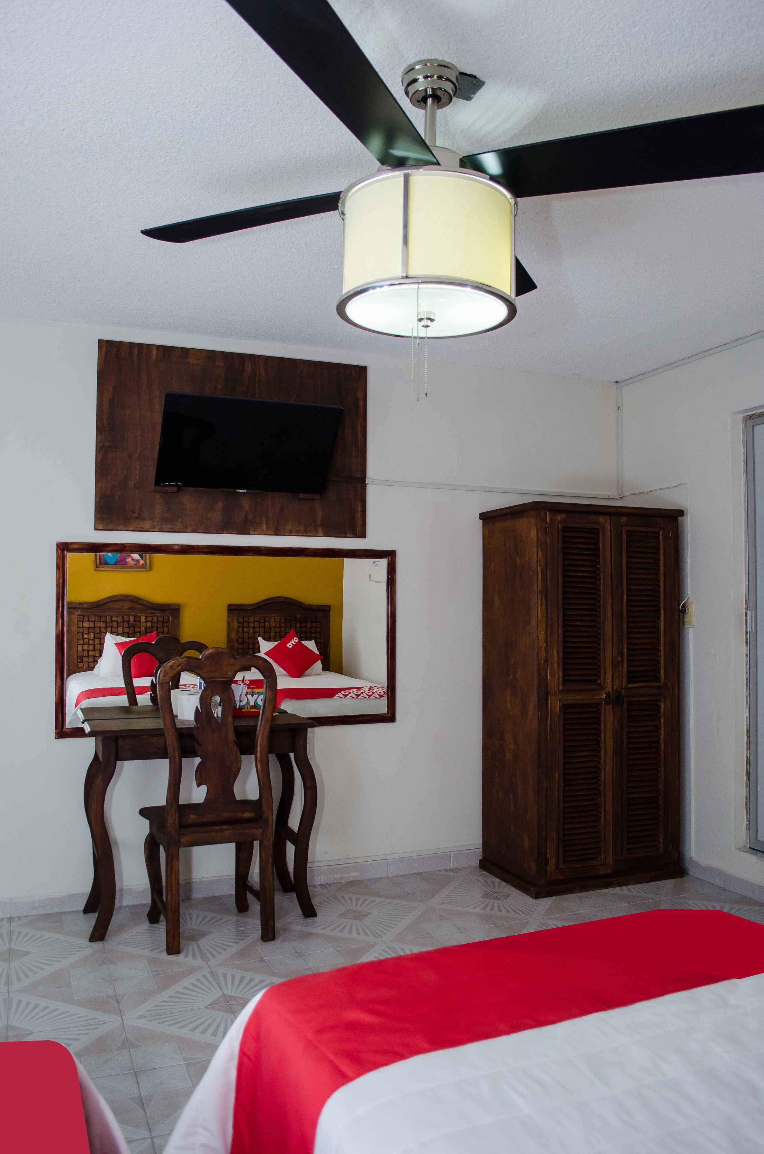 A hotel room with a bed, a wooden wardrobe, a television mounted on the wall, and a ceiling fan.