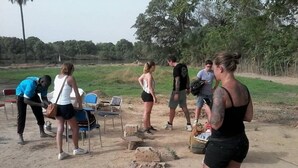 A group of people outdoors with chairs and a table, surrounded by trees and a dirt ground. - Camping Sitaba Lodge (Janjanbureh)